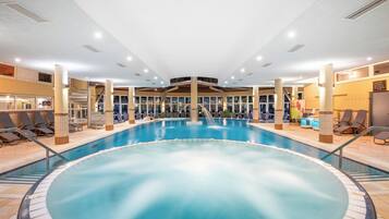 A large indoor swimming pool with a central hot tub, surrounded by lounge chairs and a staircase leading to upper levels.