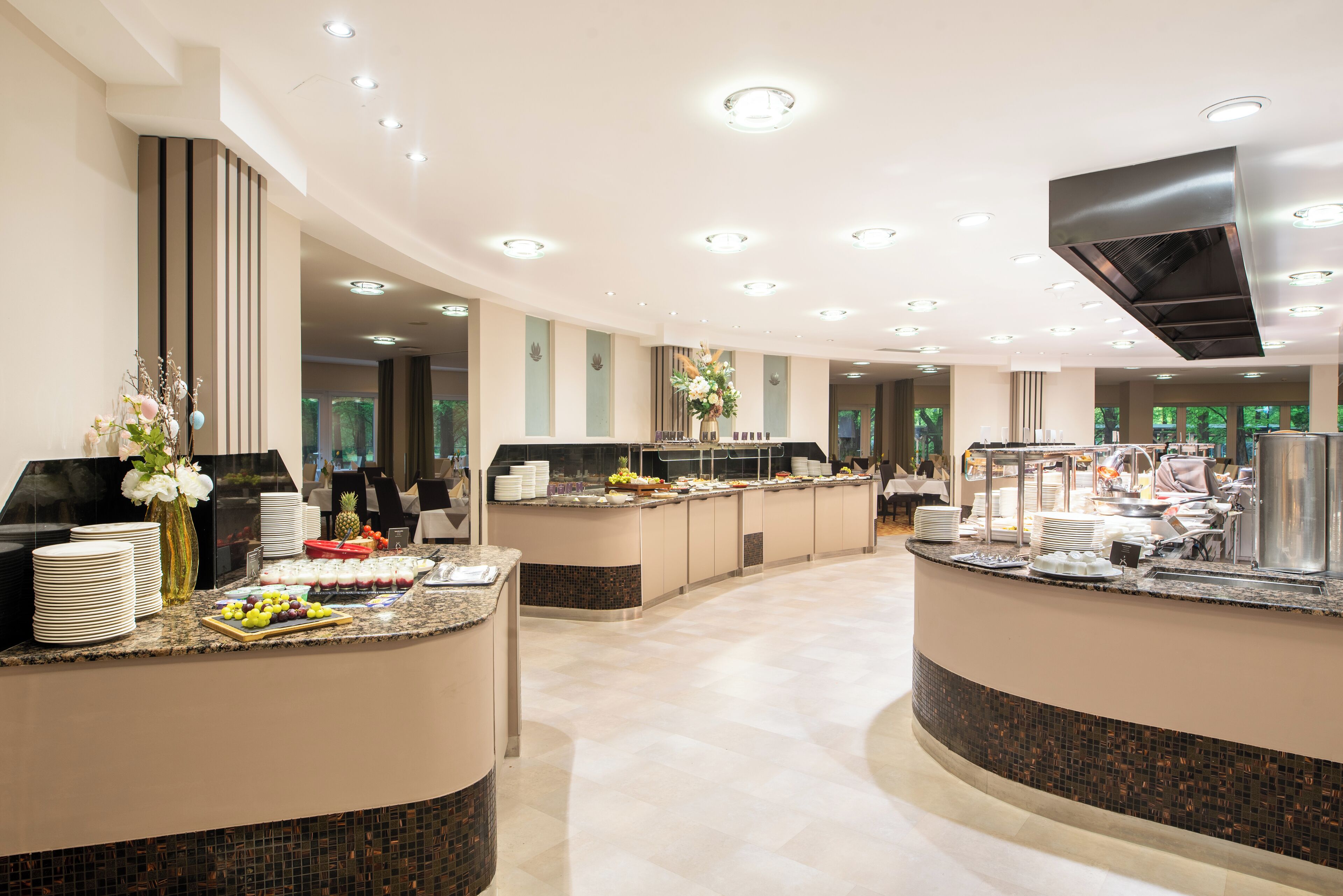 Um saguão moderno de hotel com um balcão de recepção curvo, uma tela digital grande e uma área de buffet com diversos alimentos.