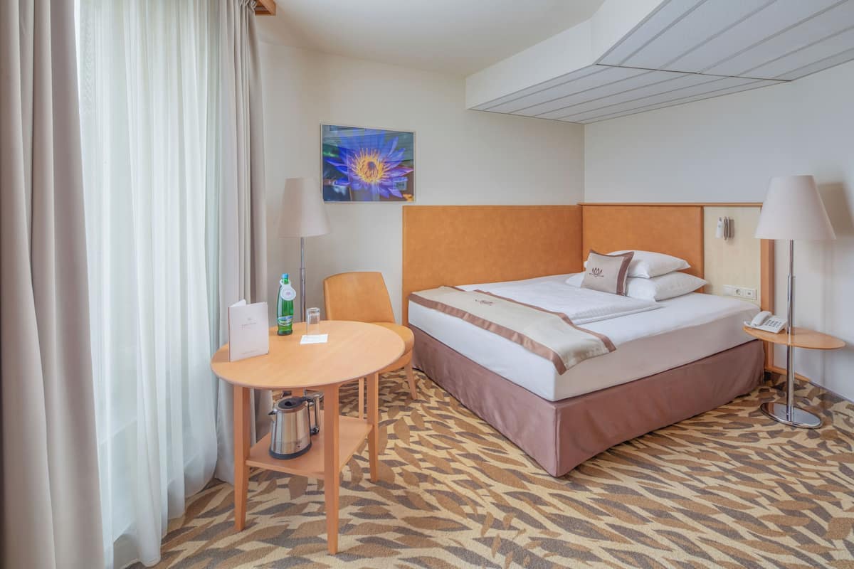 Ein Hotelzimmer mit einem Bett, einem kleinen Tisch mit Stuhl, einer Lampe, einem Bild an der Wand und einem gemusterten Teppich.