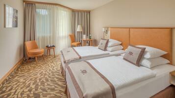 Habitación de hotel con dos camas, un área de descanso con una silla y una mesa, y un ventanal con cortinas.