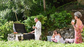 Un hombre preparando alimentos a la parrilla al aire libre, con un muro de piedra y abundante vegetación de fondo.
