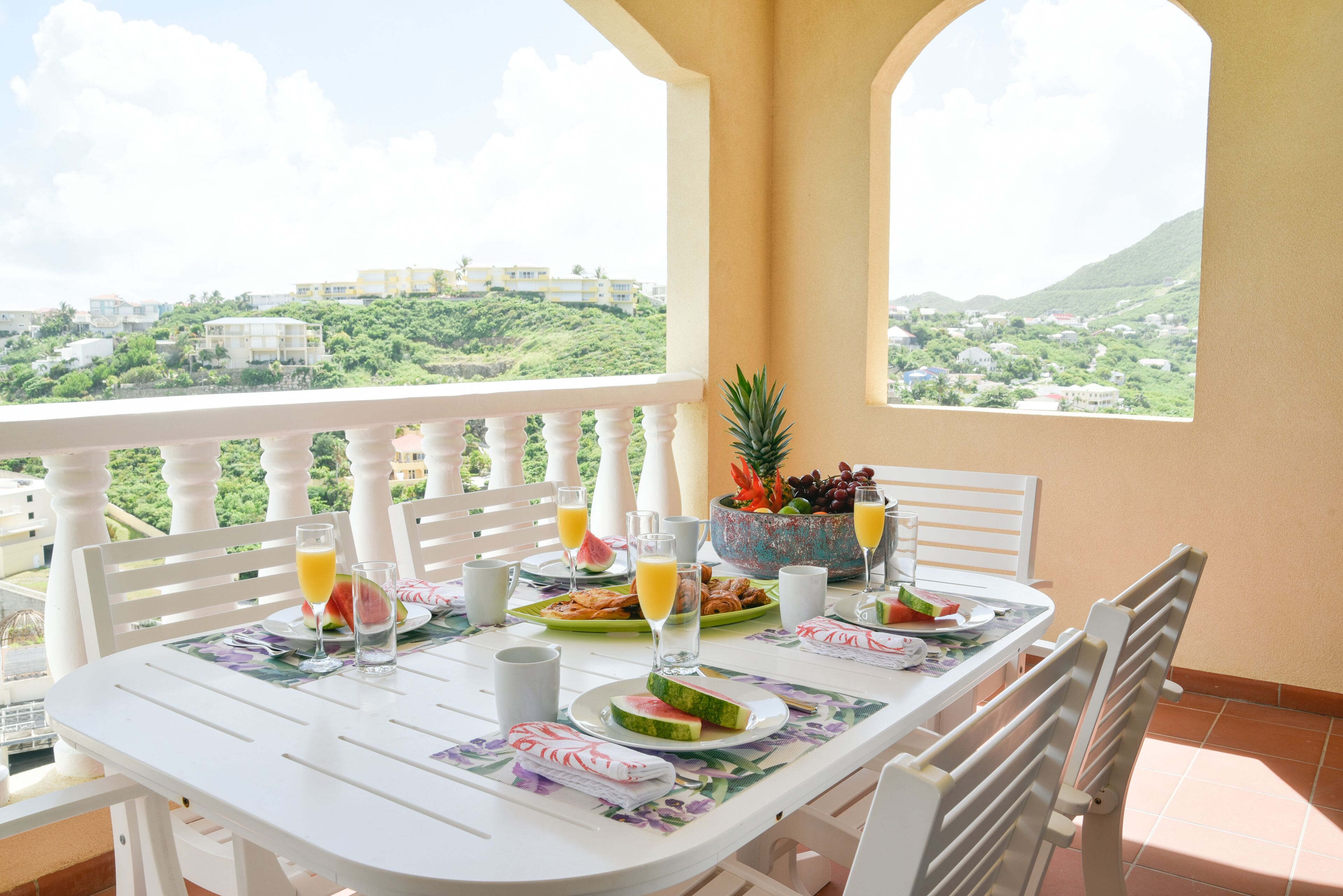 A balcony table set with breakfast items, a view of a hillside town, and a tropical fruit bowl.