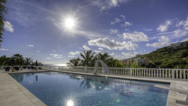 Una piscina azul cristalina con un deck blanco y palmeras, rodeada por una cerca blanca y edificios al fondo.