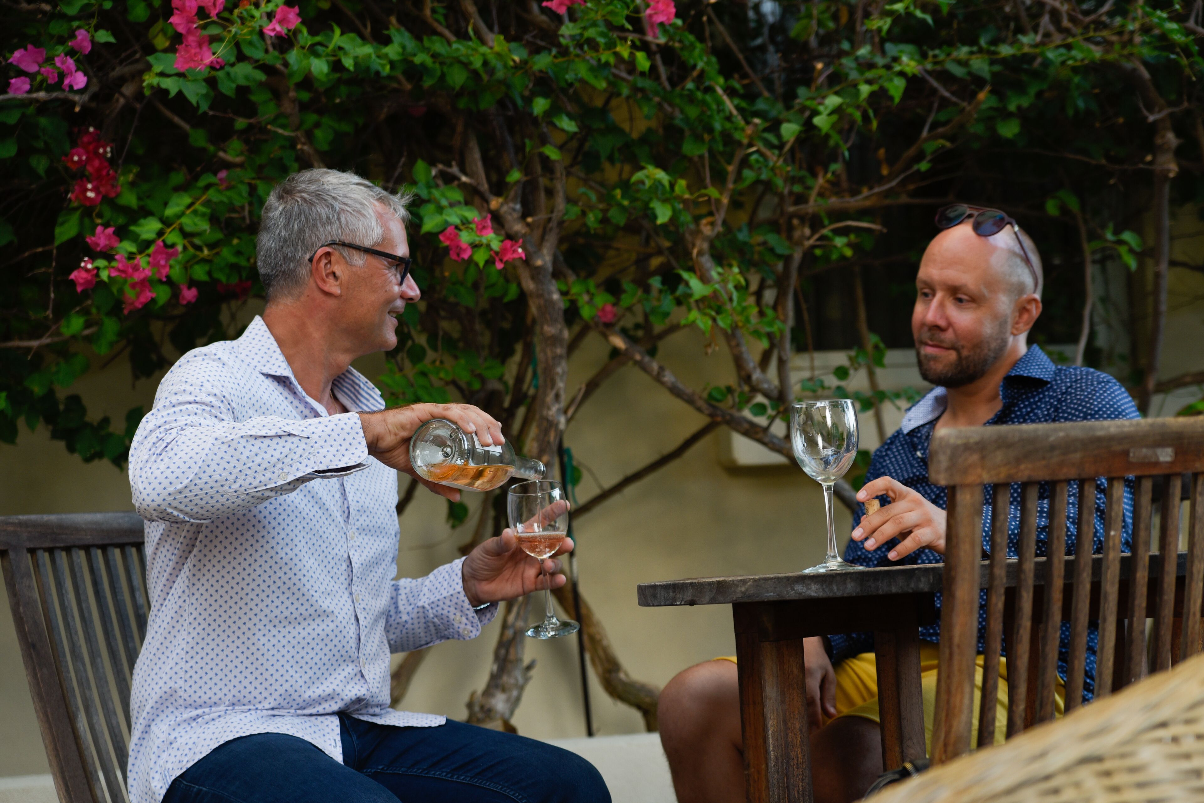 Two people sitting at a wooden table outdoors, each holding a glass of wine.