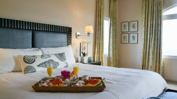A neatly made bed with a tray of breakfast items, including fruit and juice, in a hotel room with a headboard, wall art, and curtains.