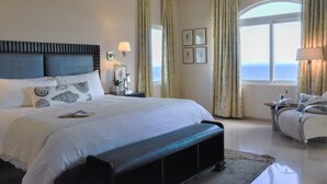 Un dormitorio con una cama grande, un banco, una silla, una mesita de noche, una lámpara y vistas al mar a través de una ventana.