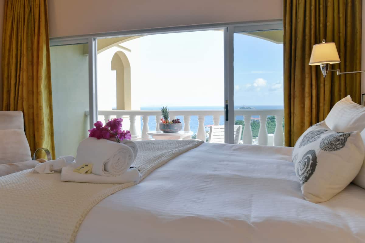 Une chambre d’hôtel avec un grand lit, un balcon donnant sur la mer et une petite table sur laquelle se trouve un panier de fruits et un vase de fleurs.