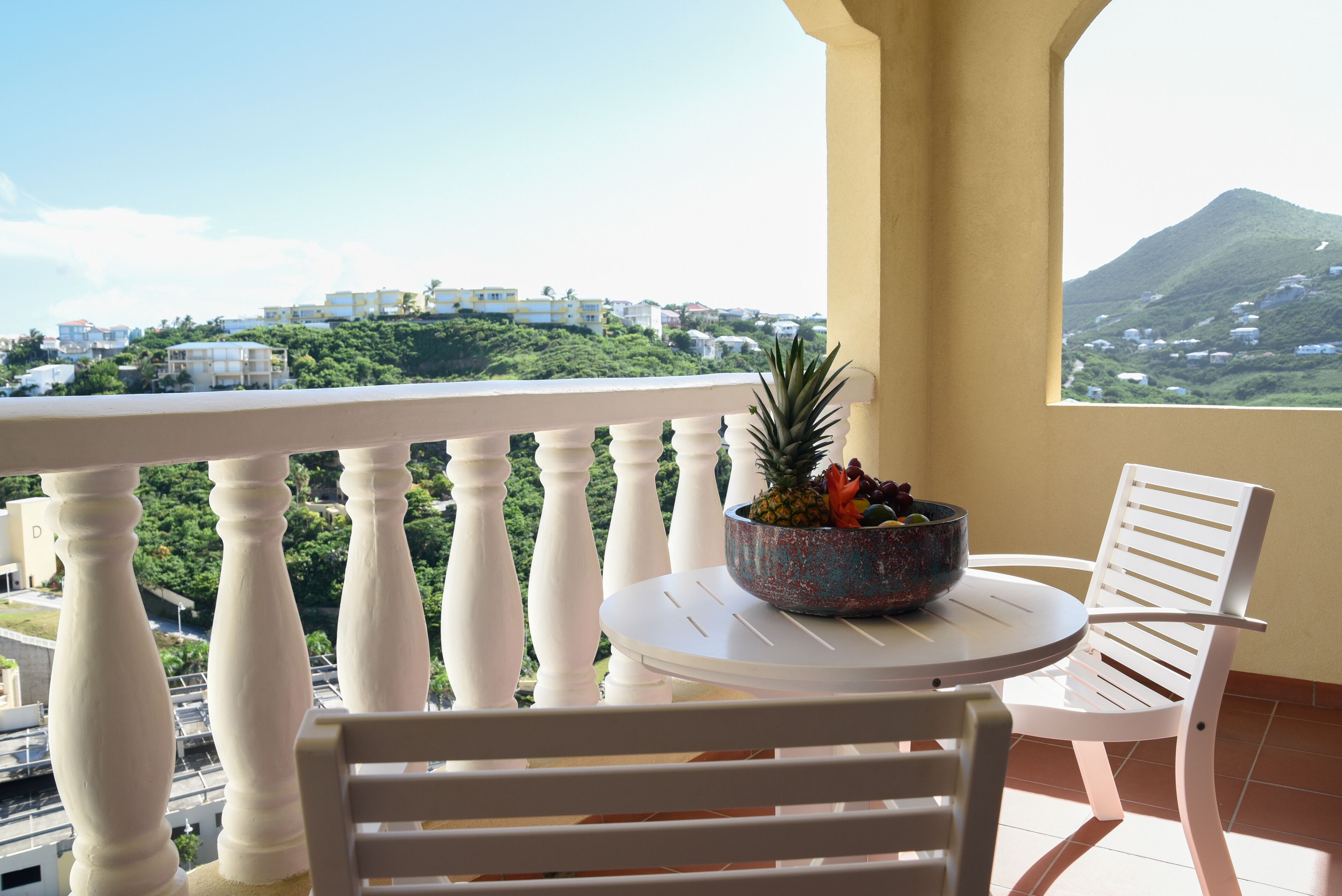 A balcony with white balustrades, a round table, and two white chairs. A bowl with fruits is on the table.