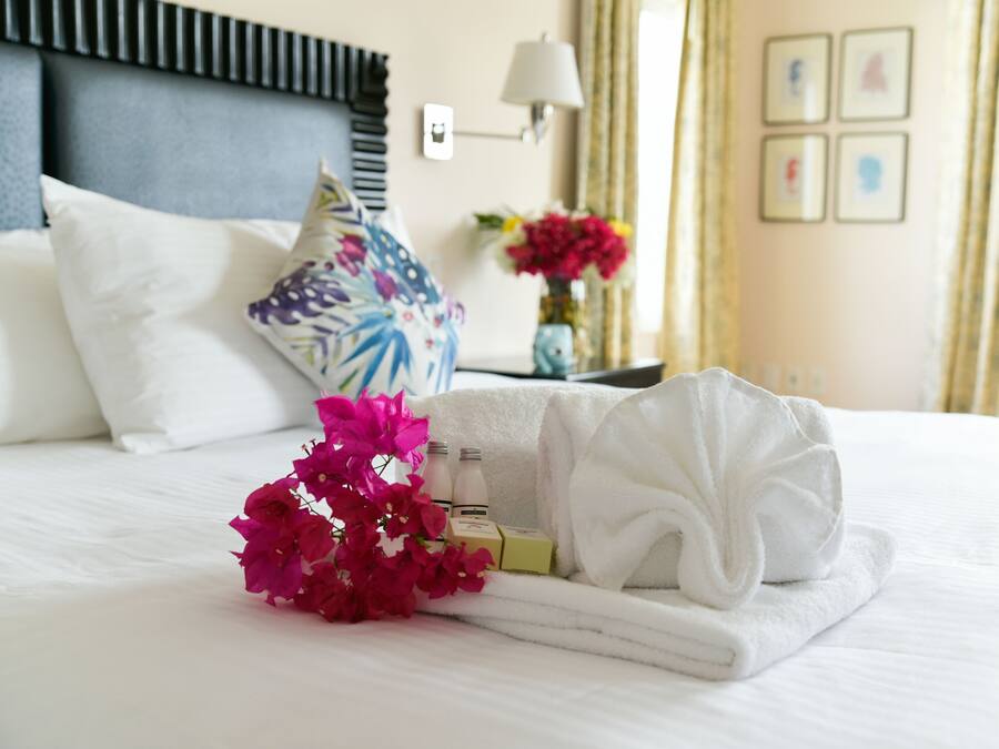A neatly made bed with a white towel, a bouquet of pink flowers, and toiletries.