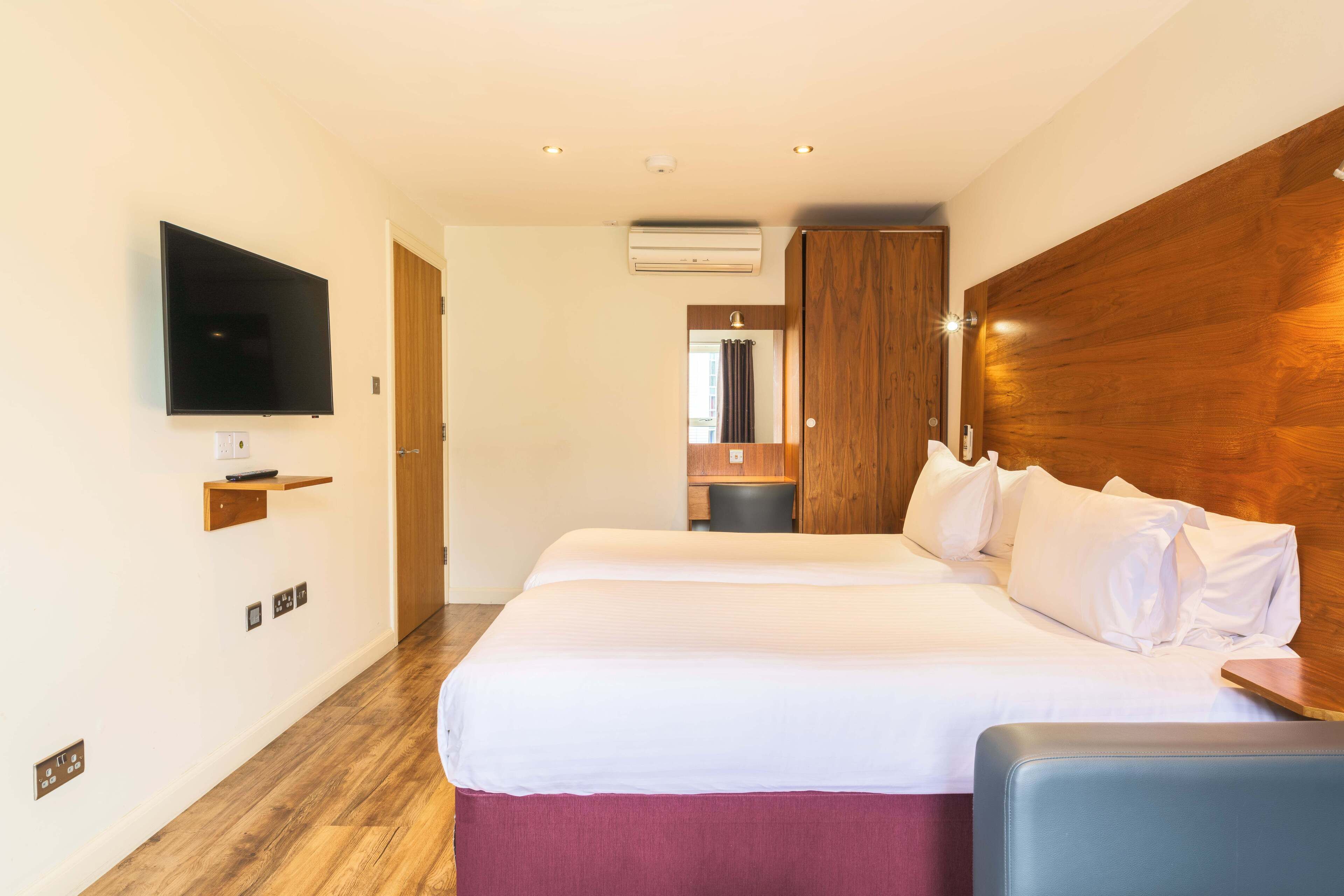 Quarto de hotel com uma cama grande, televisão presa na parede e cabeceira de madeira.