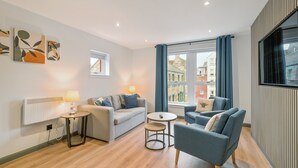 Een moderne woonkamer met een bank, fauteuil en een bijzettafeltje met een lamp. Er is een raam met gordijnen, een radiator en kunst aan de muur.