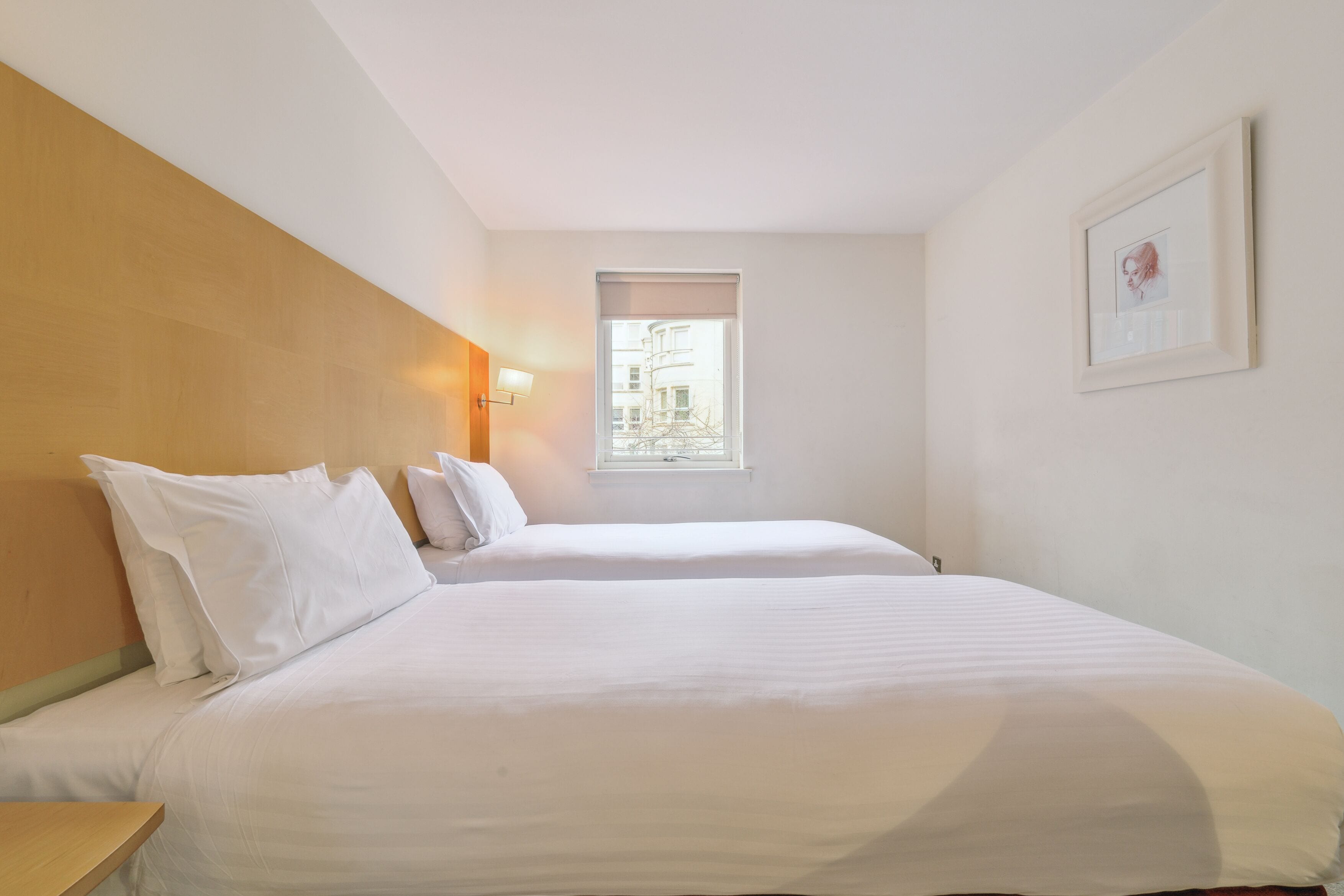 Habitació d'hotel amb dos llits individuals, una finestra amb persianes, un quadre amb marc a la paret i un panell de llit de fusta.
