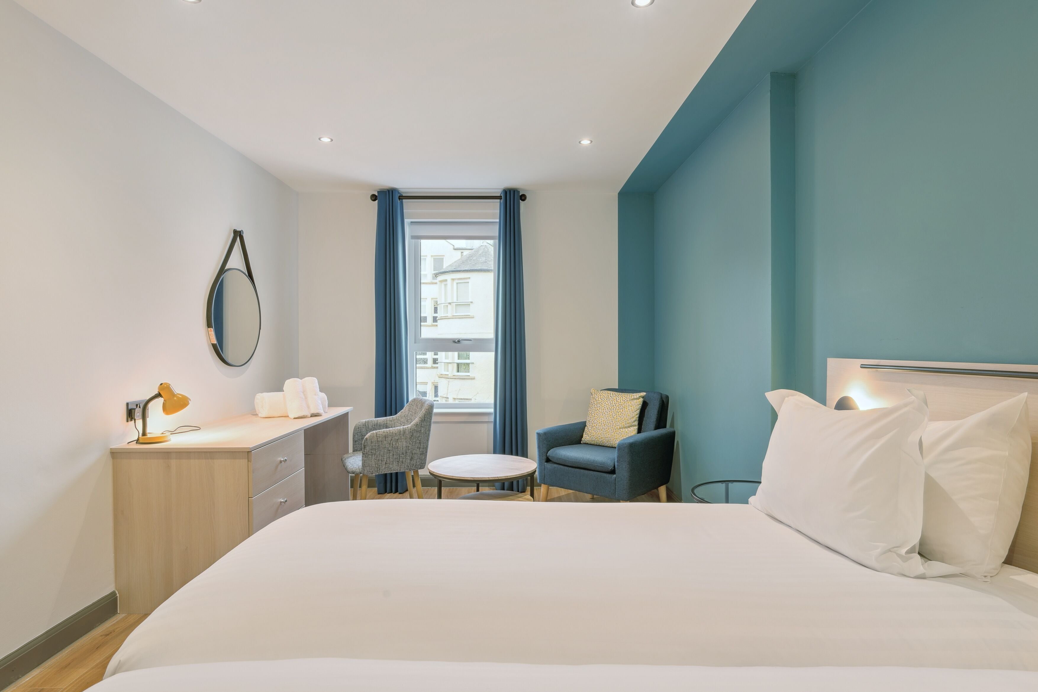 Habitación de hotel moderna con una cama grande, un escritorio con lámpara, una silla y vistas a la ciudad a través de una ventana.