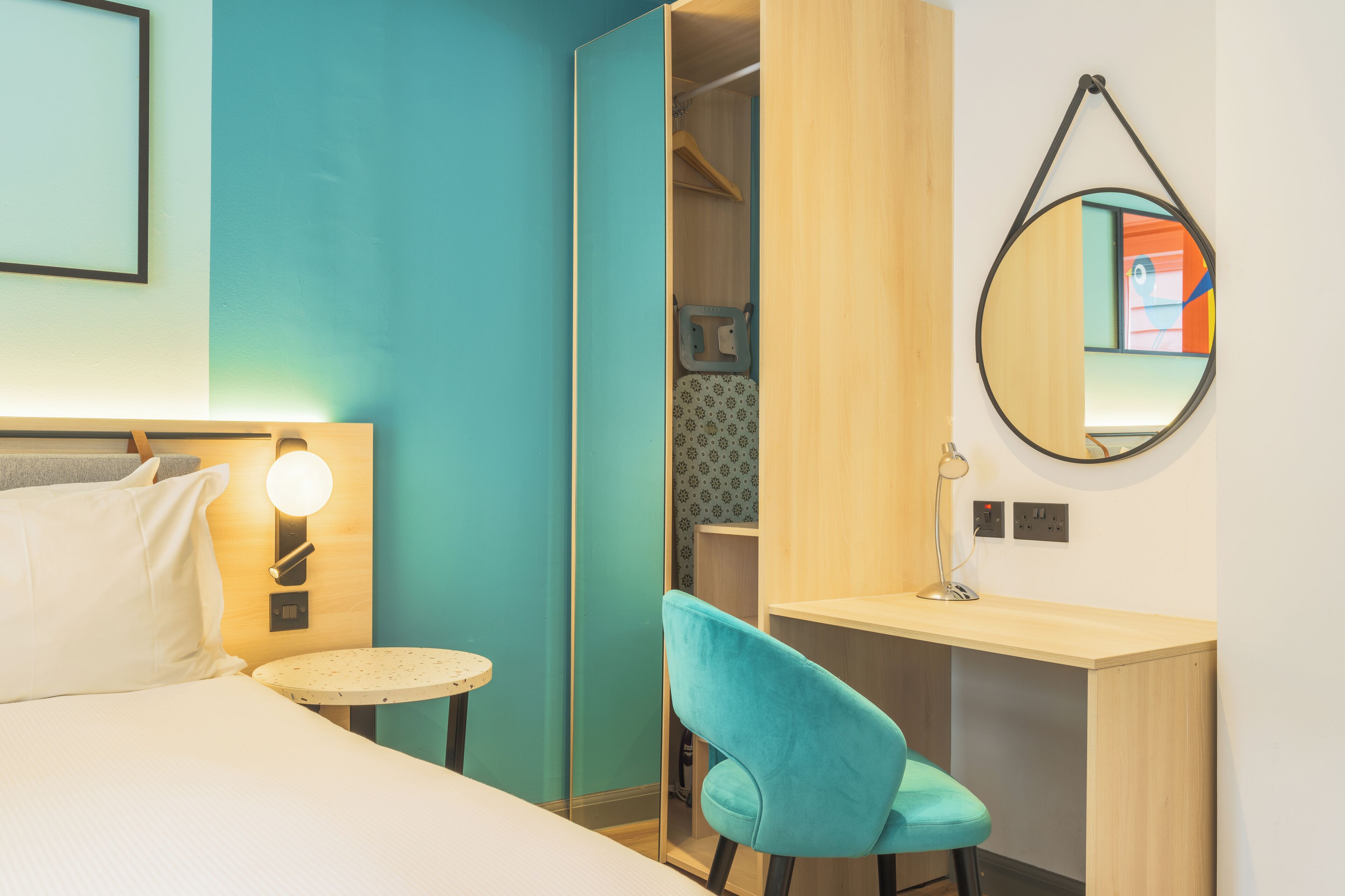 Une chambre d’hôtel avec un lit, une table ronde, une chaise bleue, un bureau, un miroir et une armoire.