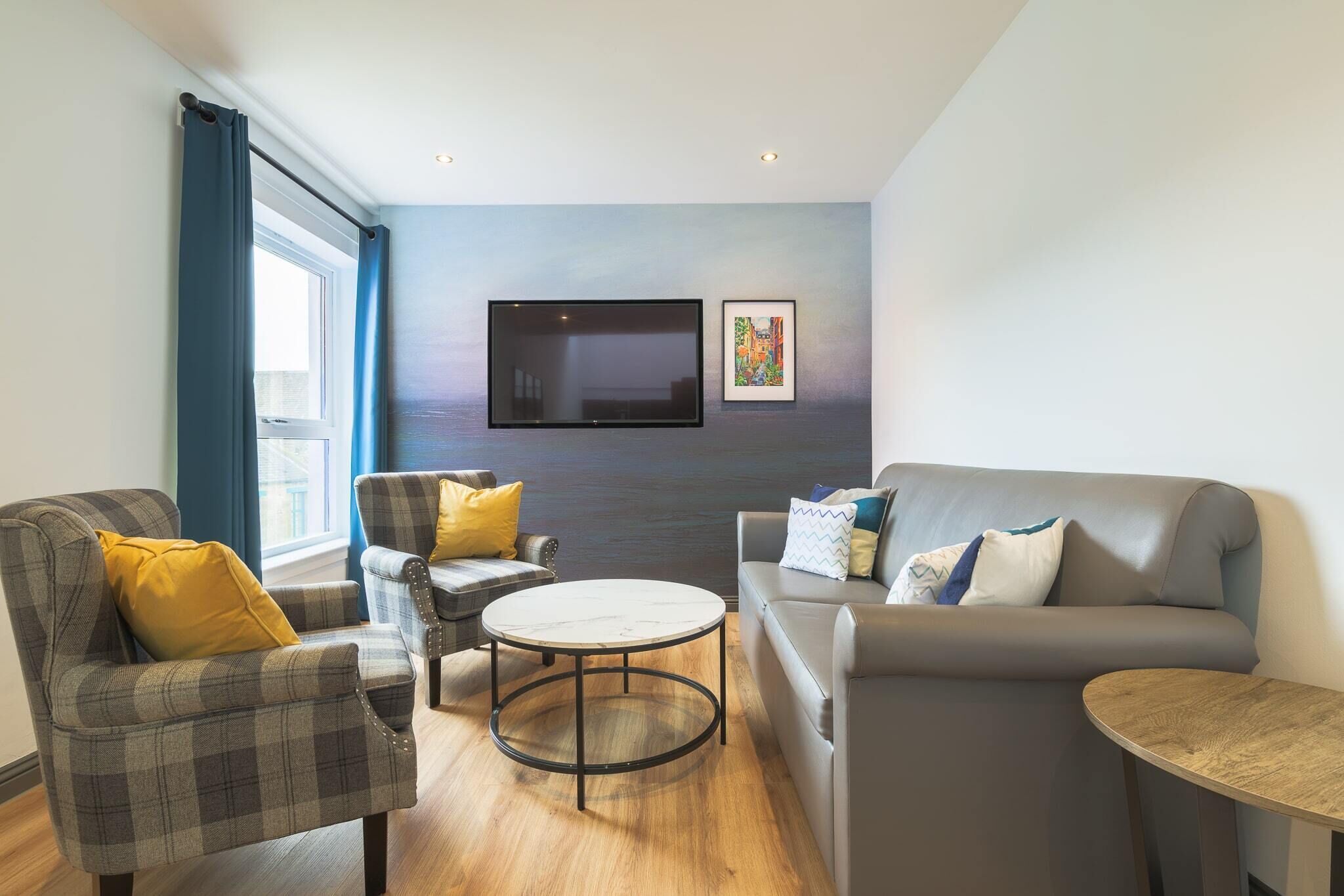 A modern living room with a grey sofa, two armchairs, a coffee table, and a flat-screen TV mounted on the wall.