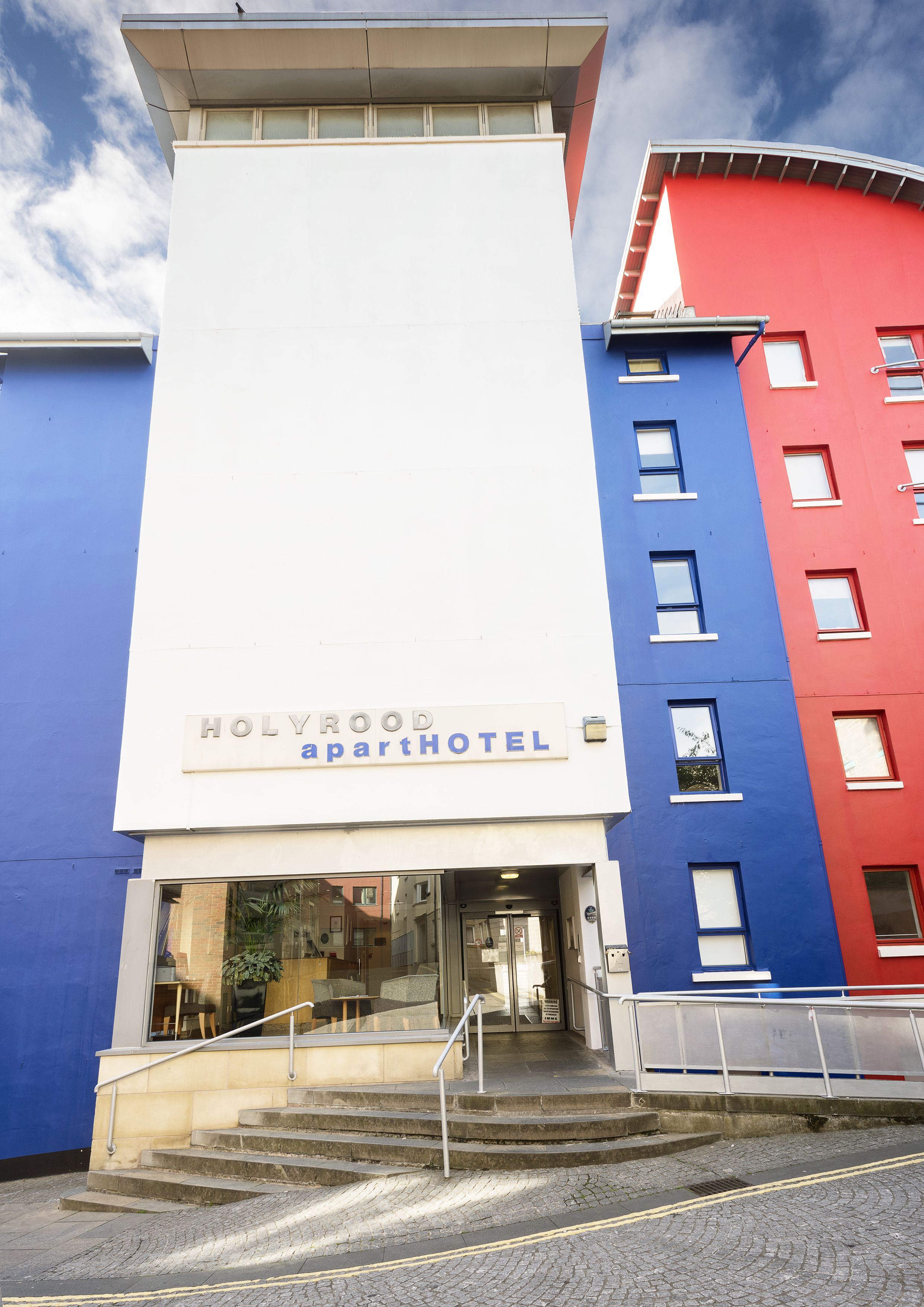 Een HOLYROOD apartHOTEL ingang met een witte gevel, een rood-blauw gebouw ernaast, en een trap die naar de ingang leidt.