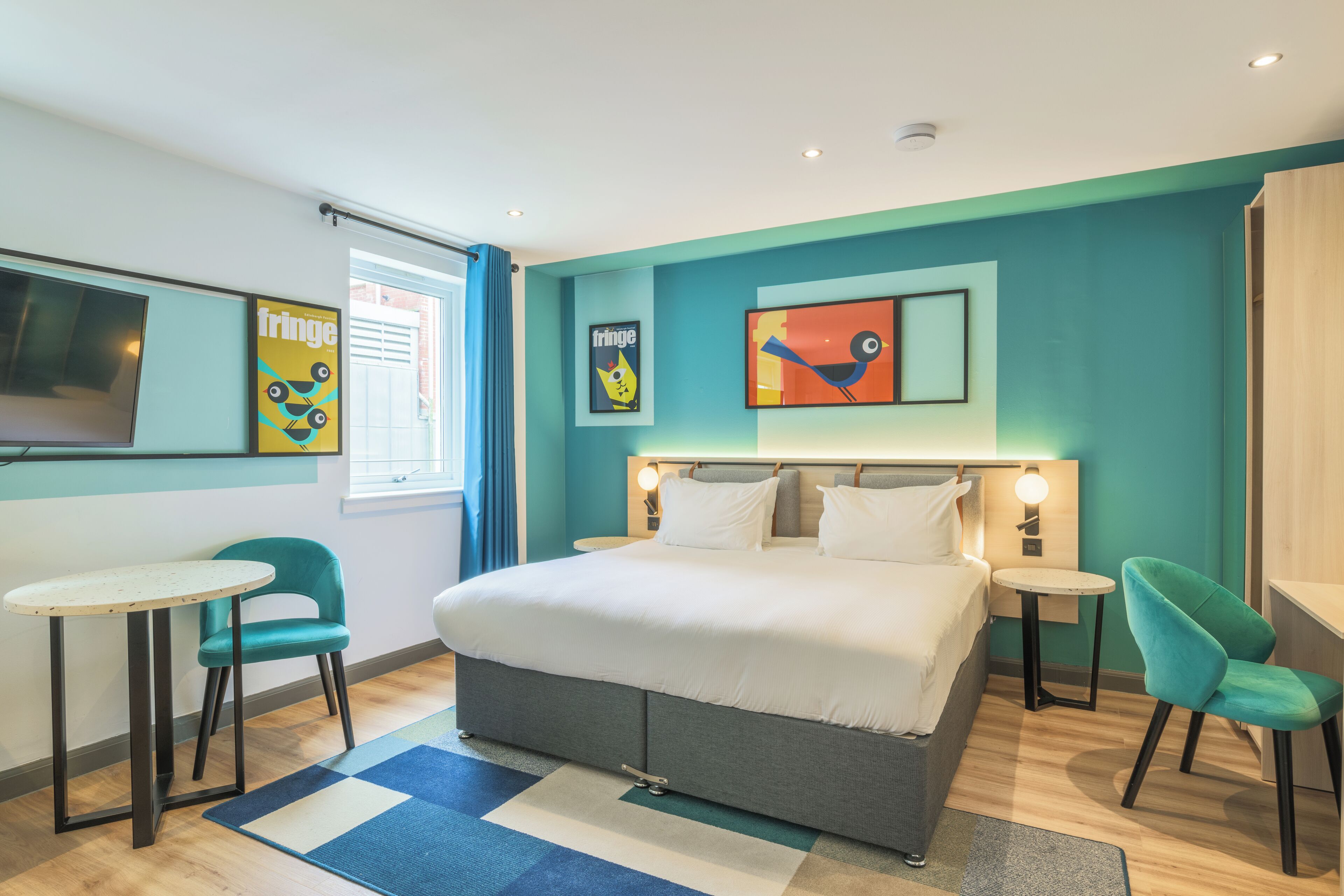 Una habitación de hotel moderna con una cama grande, dos mesitas de noche, un escritorio pequeño y dos sillas. Se observan obras de arte en la pared y una ventana con cortinas.
