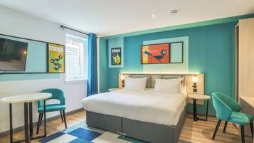 Una habitación de hotel moderna con una cama grande, dos mesitas de noche, un escritorio pequeño y dos sillas. Se observan obras de arte en la pared y una ventana con cortinas.