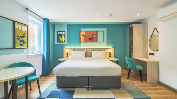 Een moderne hotelkamer met een groot bed, een bureau en een stoel. Er hangen twee schilderijen aan de muur, een raam met gordijnen en een klein tafeltje.