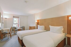 A hotel room with two beds, a desk, chairs, and a window.