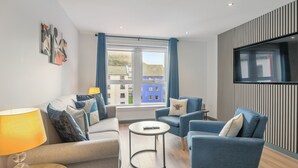 Een moderne woonkamer met een bank, fauteuils, een salontafel en een aan de muur bevestigde televisie.