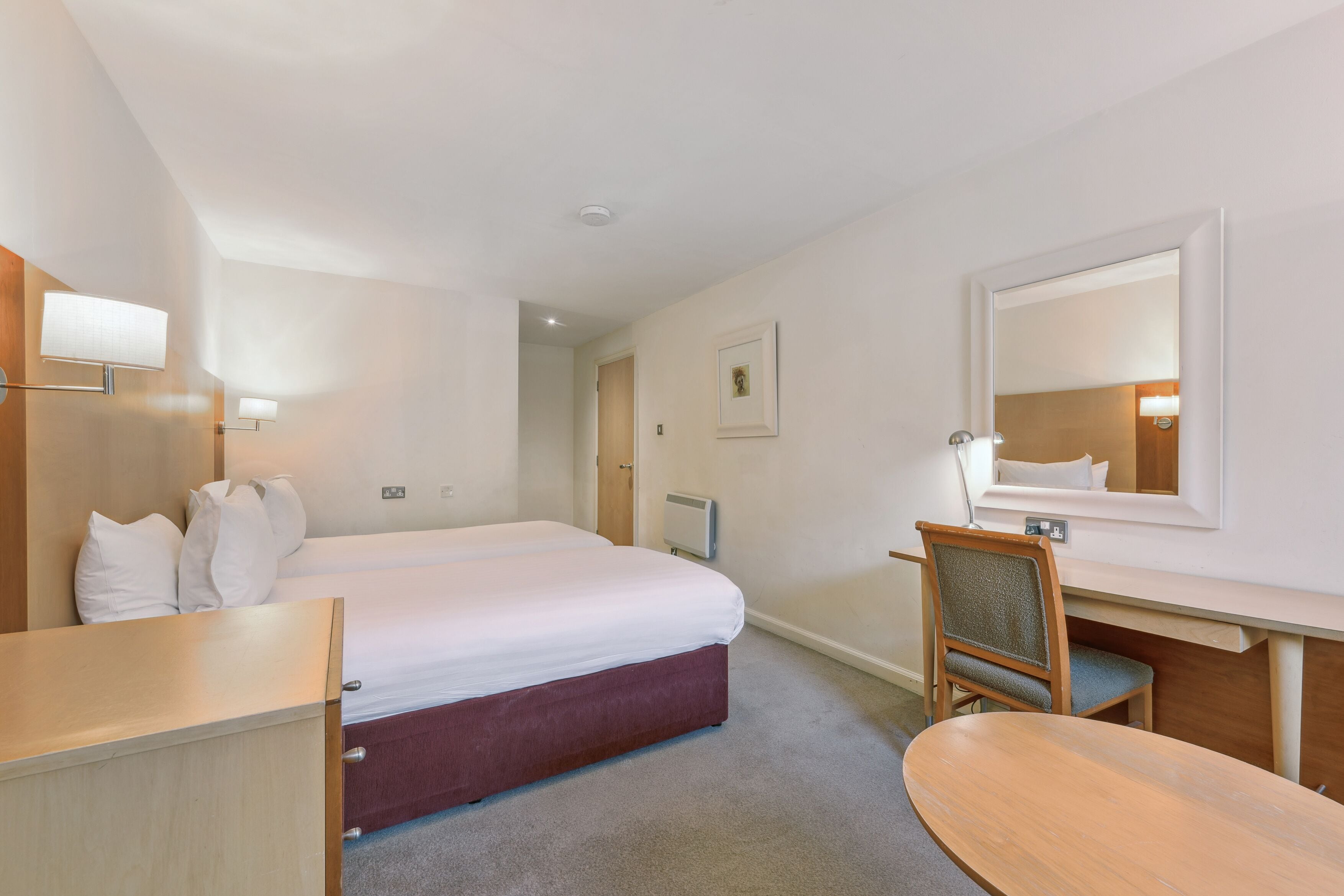 A hotel room with a bed, a desk, a chair, a mirror, and a picture on the wall.