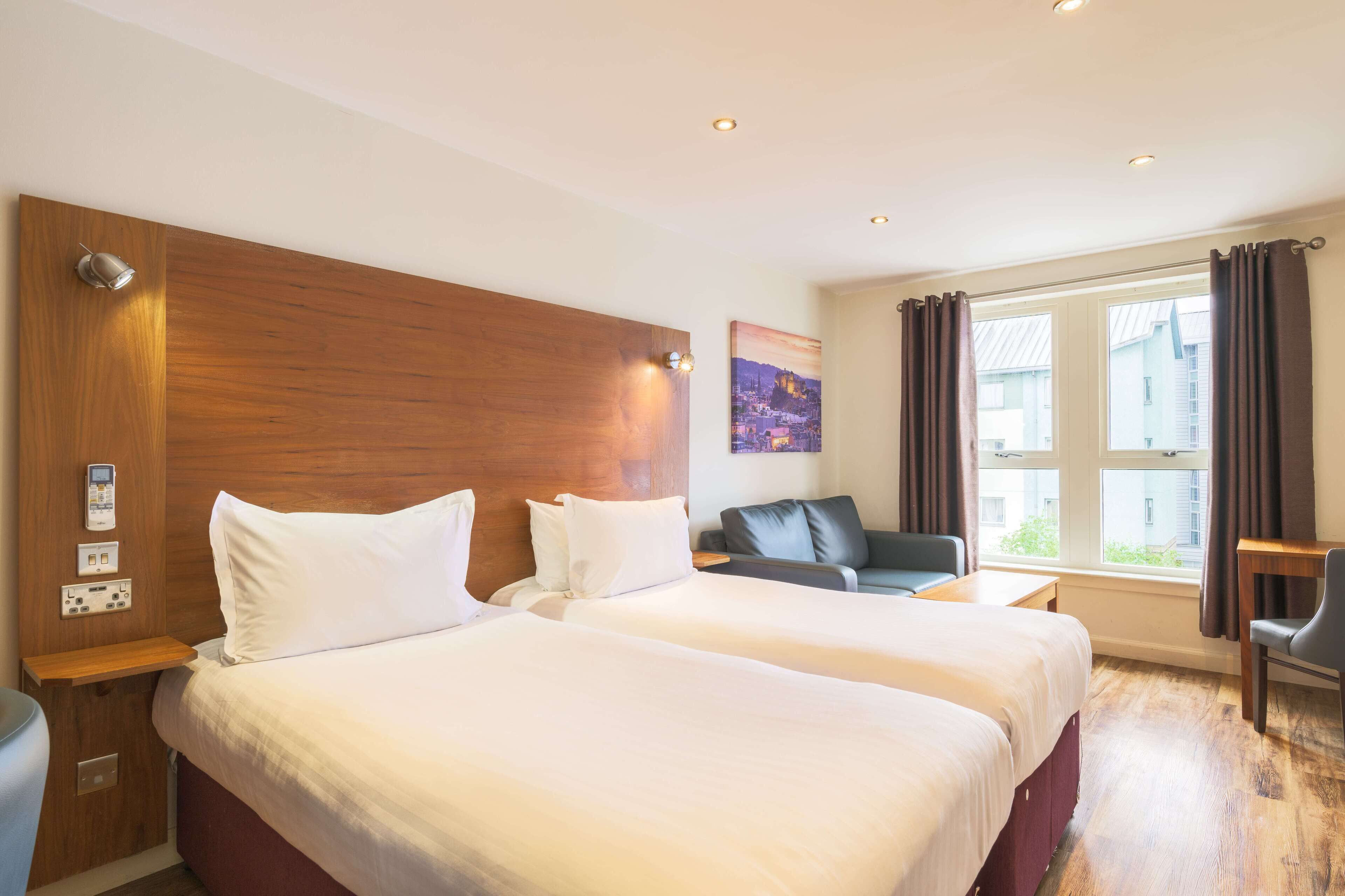 A hotel room with two beds, a desk, a chair, a sofa, and a window with curtains.