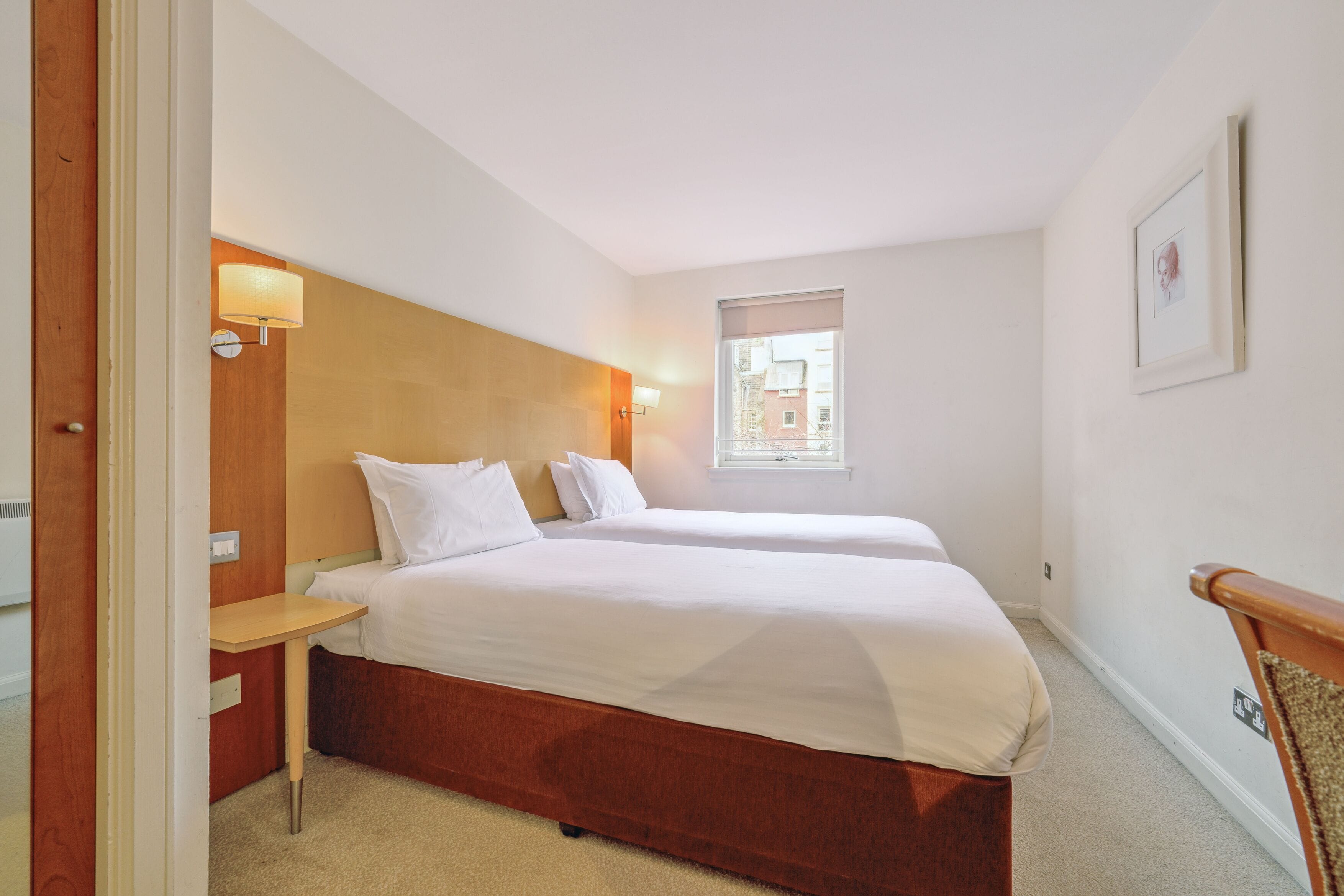 A hotel room with two beds, a wooden headboard, a nightstand, a wall-mounted lamp, a window, and a framed picture on the wall.