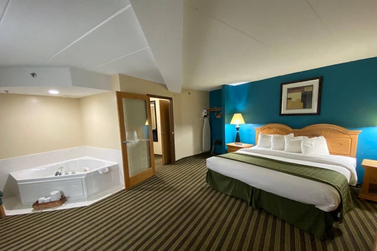A hotel room with a large bathtub, a bed with a green bedspread, a wooden headboard, and a blue accent wall.