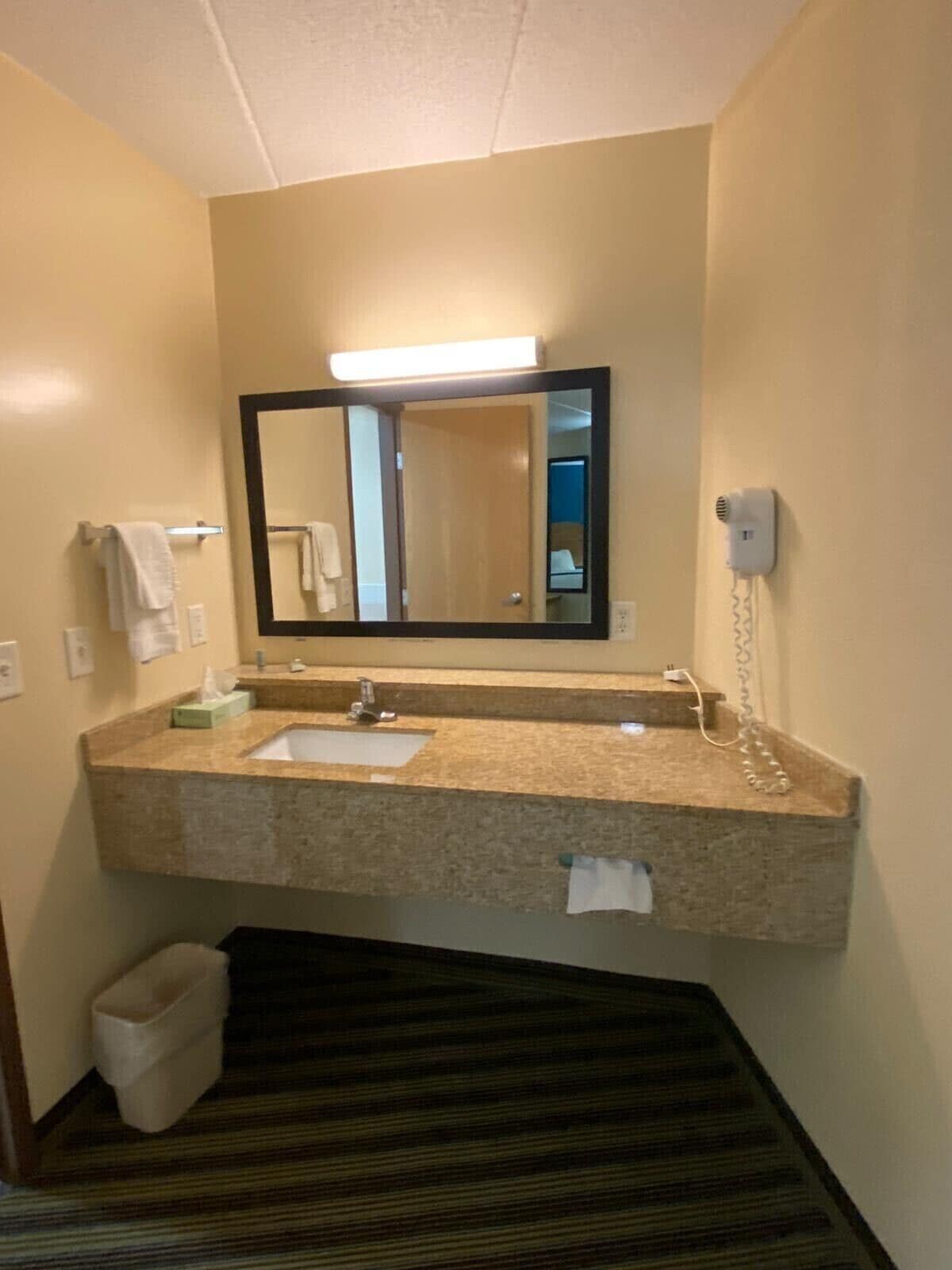 A hotel bathroom with a large mirror, two sinks, a towel rack, a trash bin, and a hairdryer on the wall.