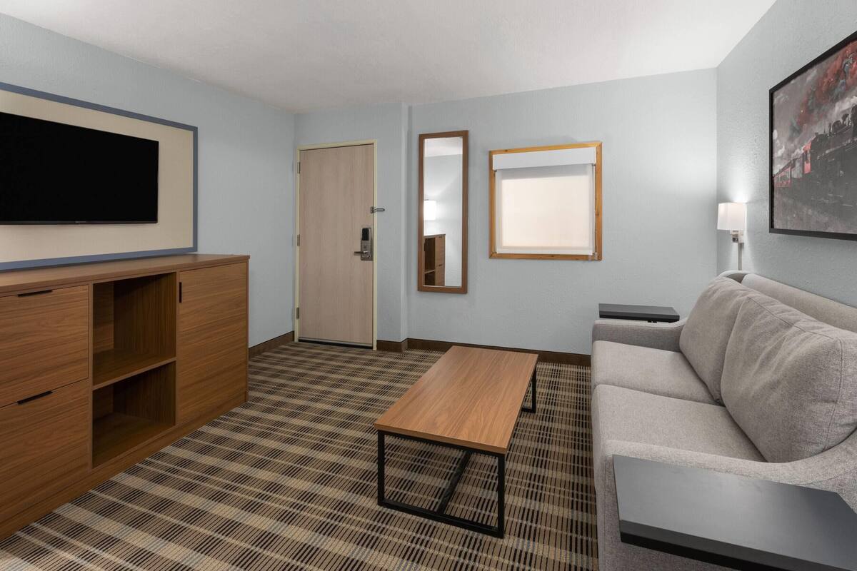 Une chambre d’hôtel équipée d’un téléviseur à écran plat, d’une armoire en bois, d’un canapé, d’une table basse, d’une porte, d’un miroir et d’un tableau accroché au mur.