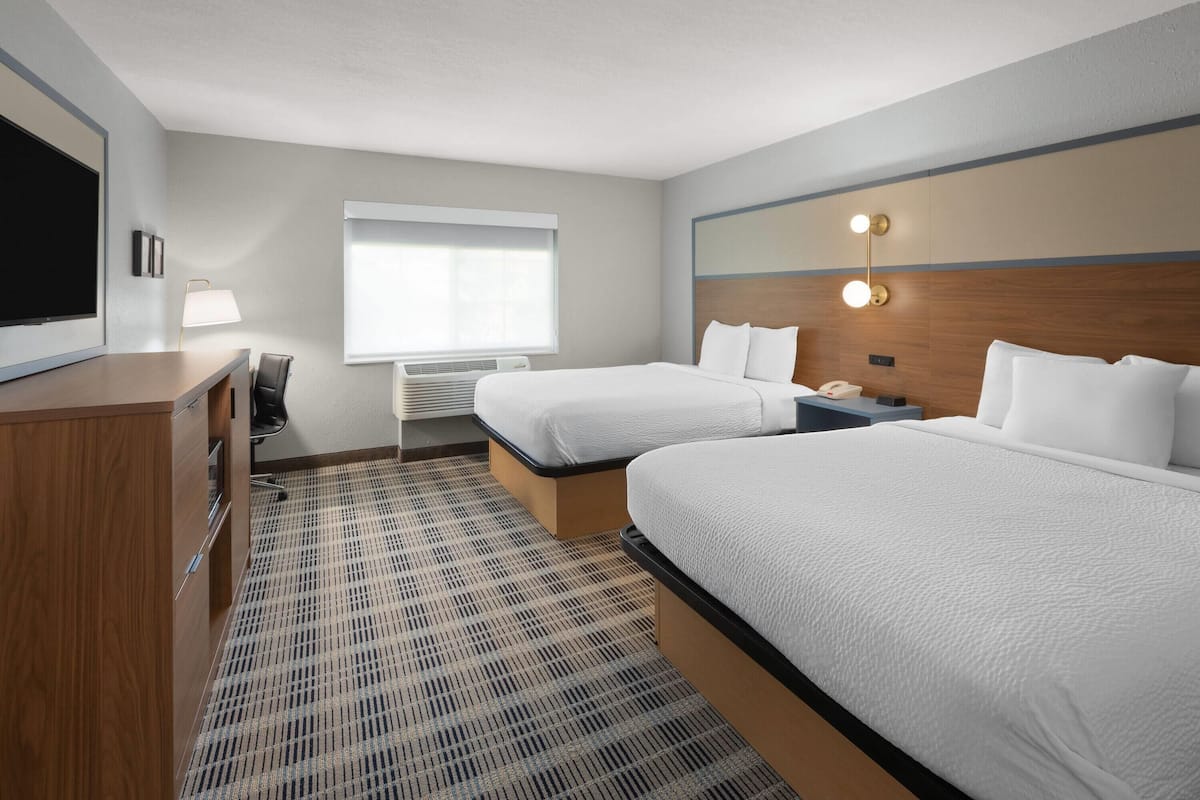 Une chambre d’hôtel avec deux lits, un bureau, une chaise, une télévision et une fenêtre avec des stores.