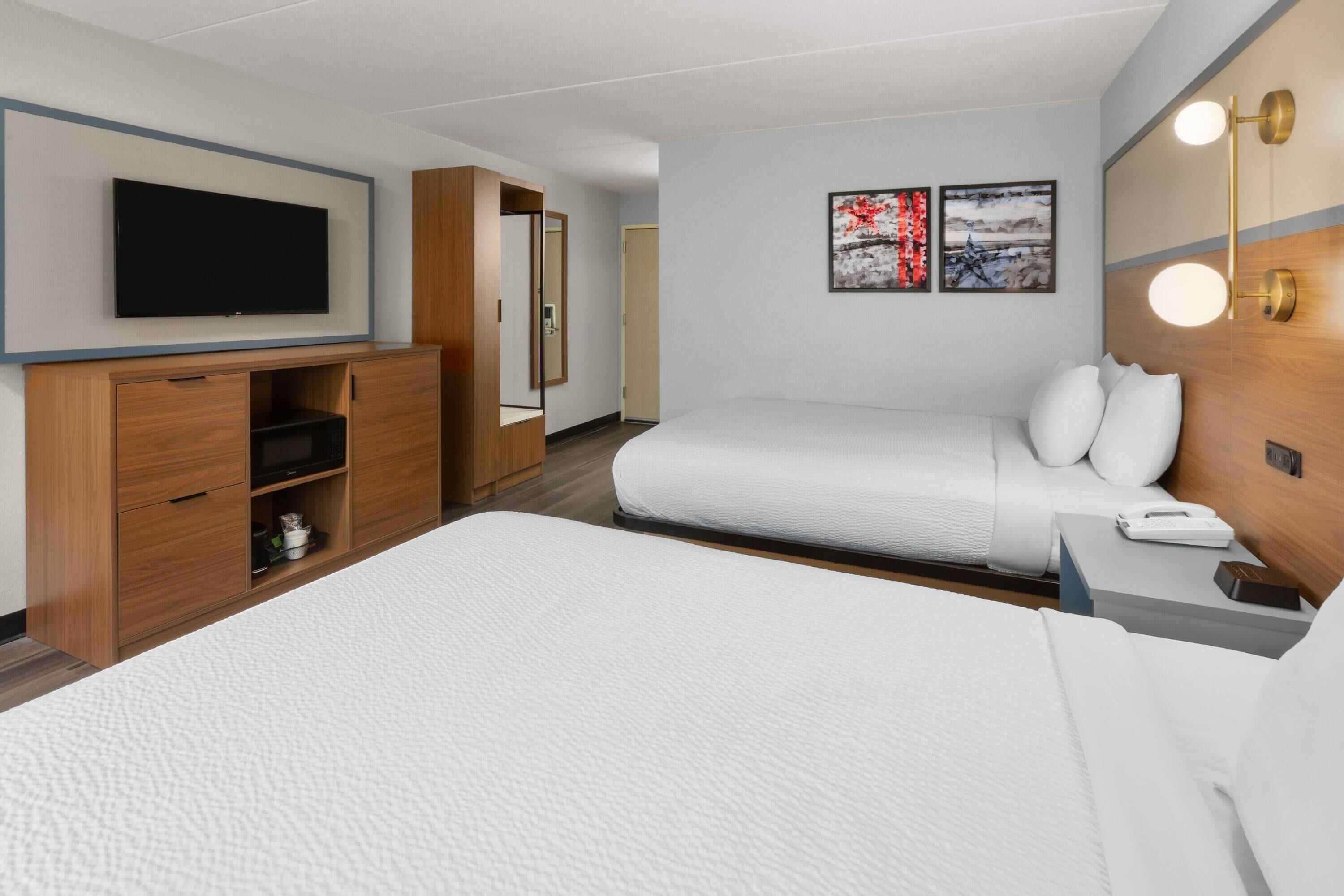 A hotel room with a large bed, a TV mounted on the wall, a wooden wardrobe, and a bedside table with a lamp.