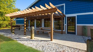 Un espace extérieur aménagé, abrité et doté de bancs et d’une pergola, situé à côté d’un bâtiment avec de grandes fenêtres et une façade bleue.