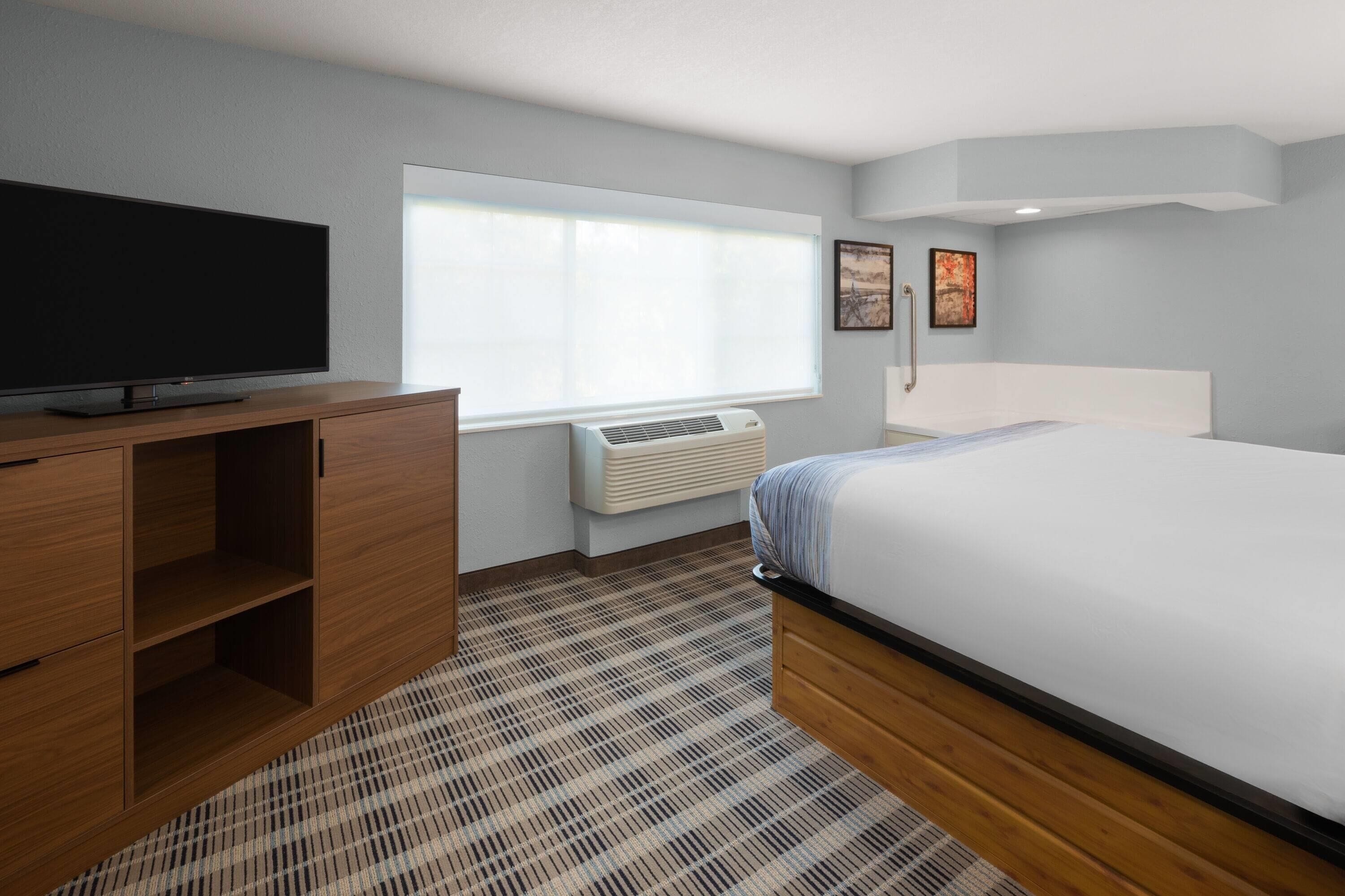 Une chambre d’hôtel avec un lit, un téléviseur posé sur une commode en bois, une fenêtre avec des stores et un climatiseur fixé au mur.
