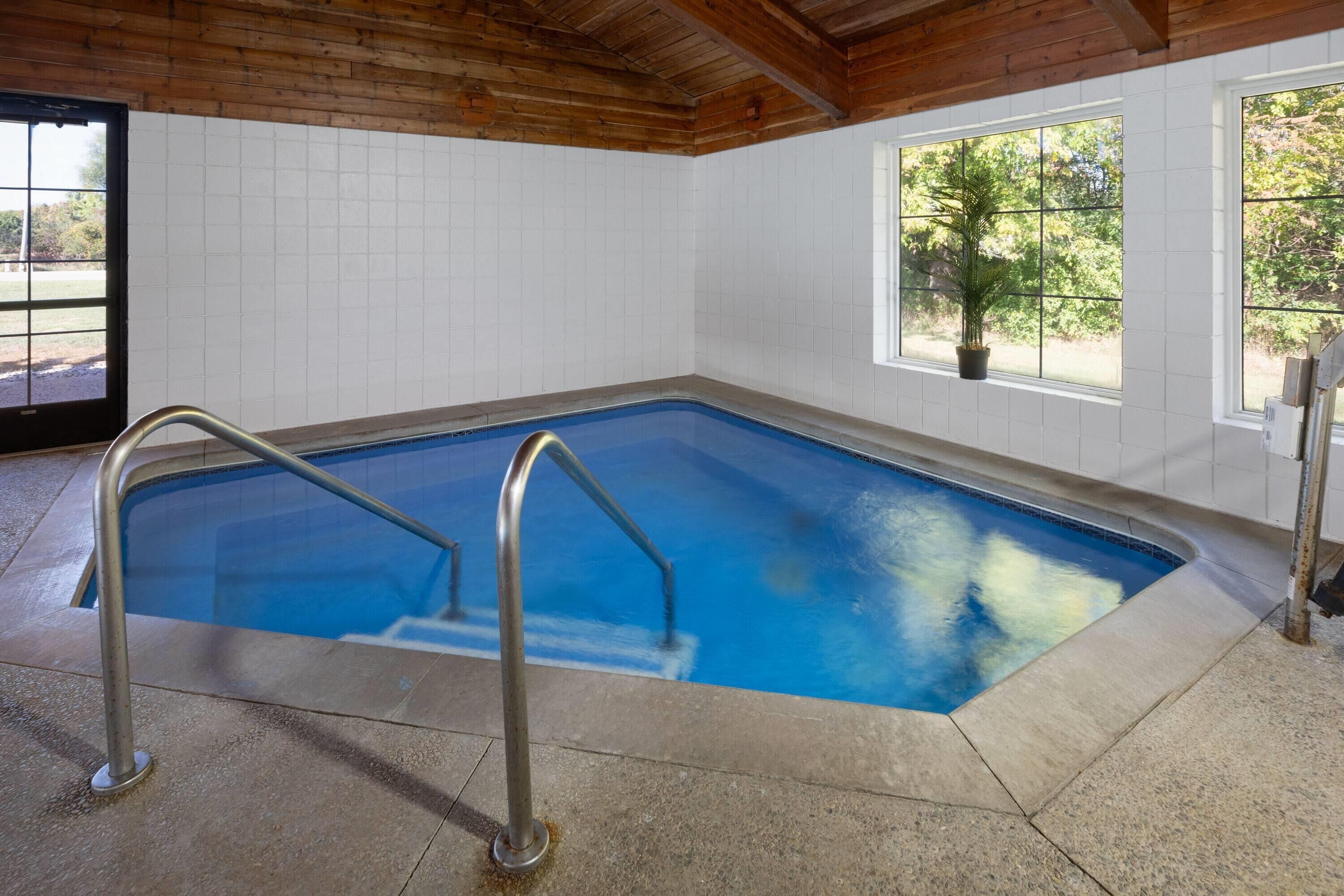 An indoor swimming pool with a wooden ceiling, tiled walls, and windows with a view of greenery.