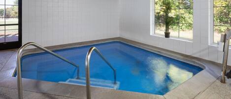 Una piscina interior con techo de madera, paredes con azulejos y ventanas con vistas a zonas verdes.
