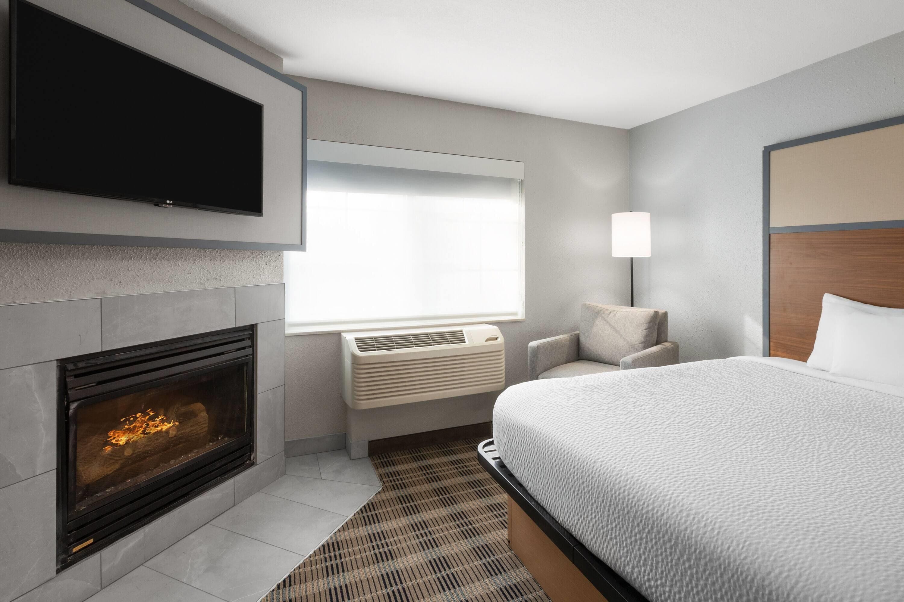 A modern hotel room with a fireplace, a flat-screen TV, a bed, and a window with blinds.