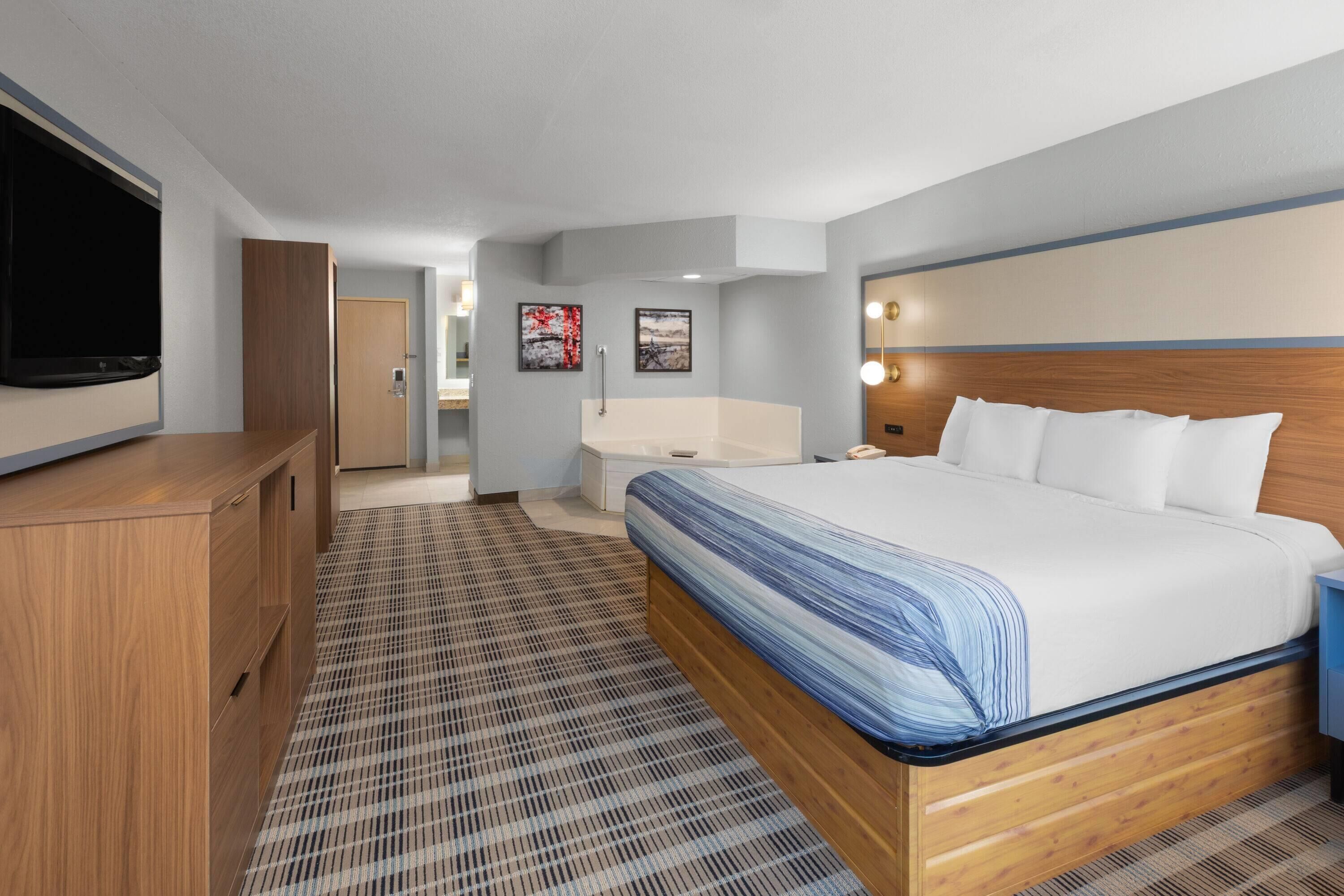 Quarto de hotel com uma cama grande, televisão, cómoda de madeira e casa de banho visível através de uma porta aberta.
