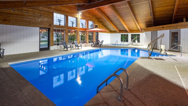Une piscine intérieure à l’eau d’un bleu cristallin, avec des poutres en bois au plafond et un espace détente comprenant des chaises et une table.