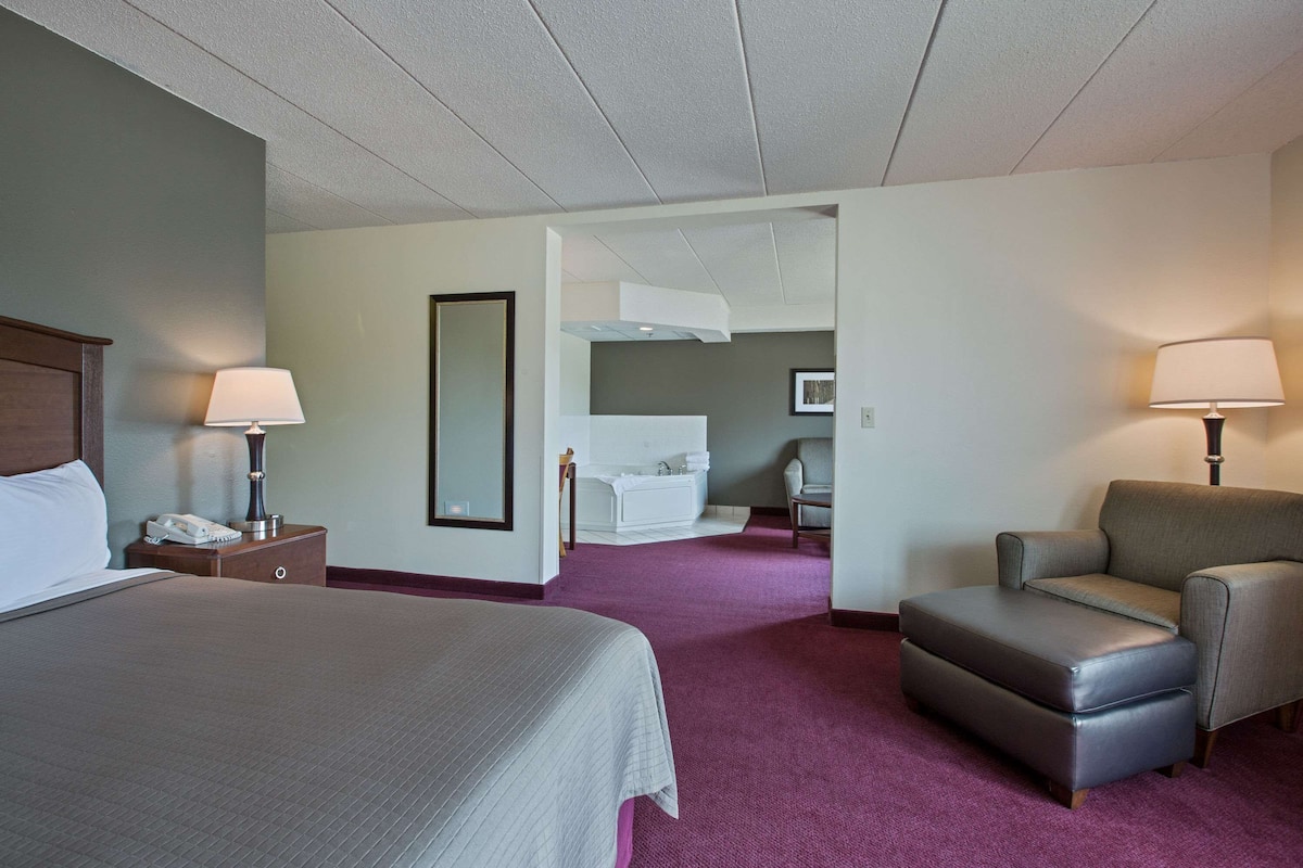 A hotel room with a large bed, a chair, a desk, a lamp, and a bathroom visible through an open door.