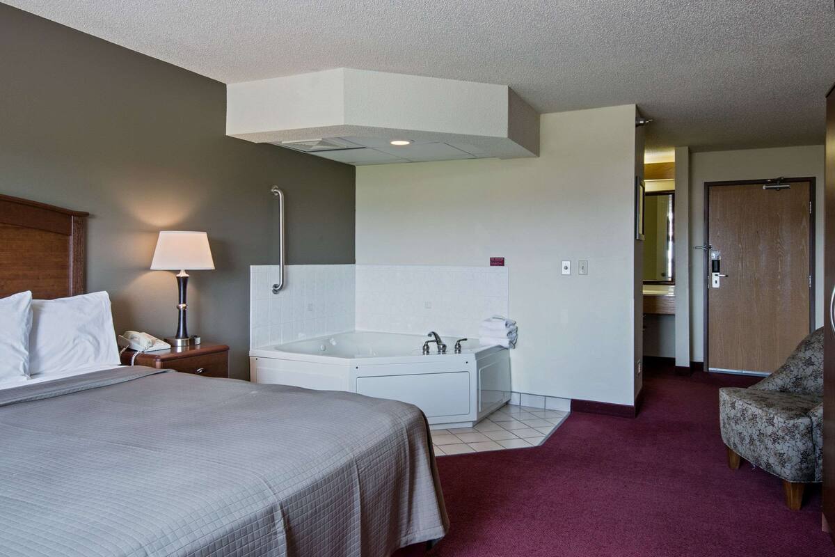 A hotel room with a large bed, a separate bathroom with a bathtub, a chair, and a desk with a lamp.