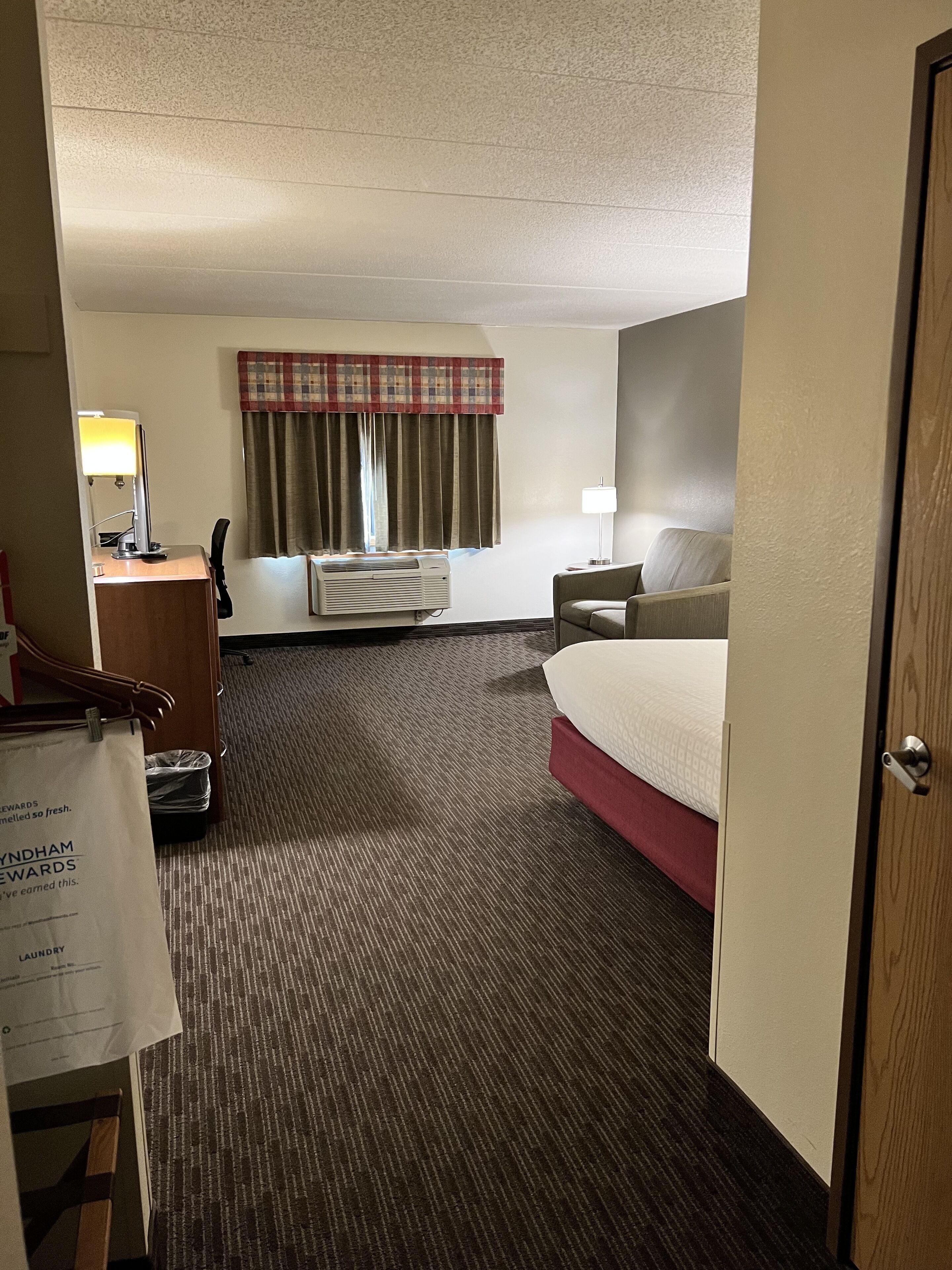 Habitación de hotel con cama, escritorio con lámpara, una silla, ventana con cortinas y piso alfombrado.