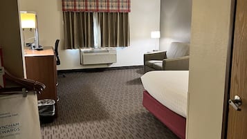 Habitación de hotel con cama, escritorio con lámpara, una silla, ventana con cortinas y piso alfombrado.