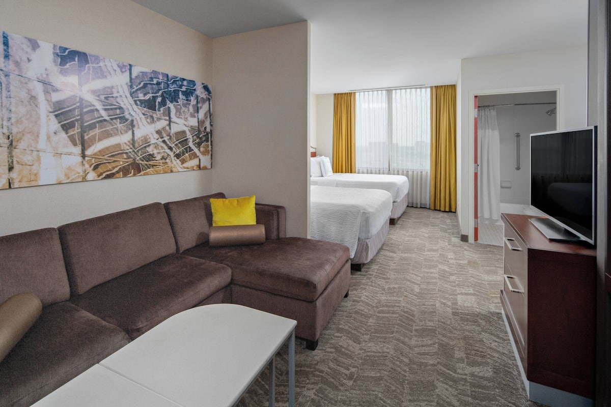 A hotel room with a brown sofa, a white table, a TV on a wooden cabinet, and a bed with white linens.