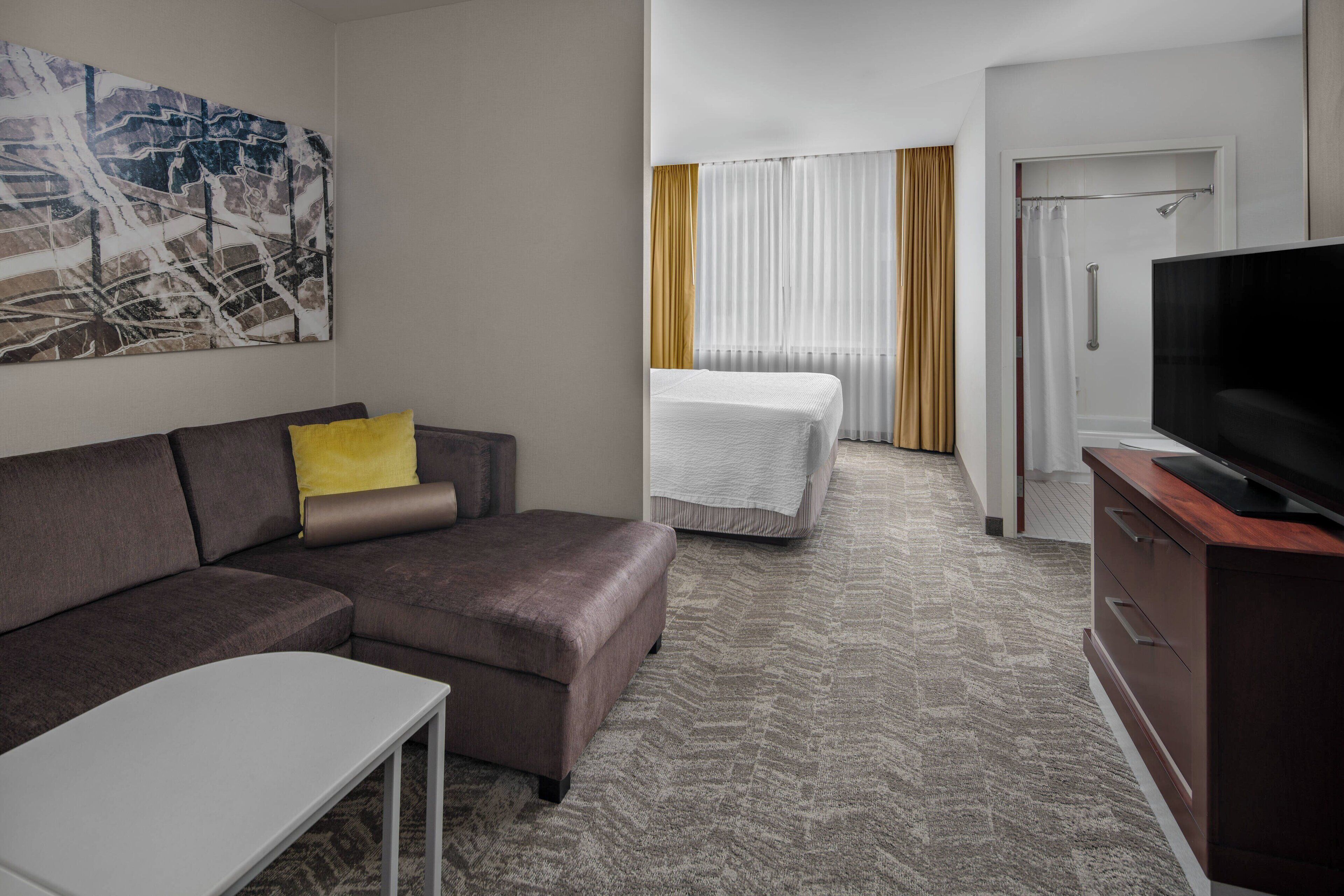 A hotel room with a brown sofa, a white bed, a TV, and a painting on the wall.