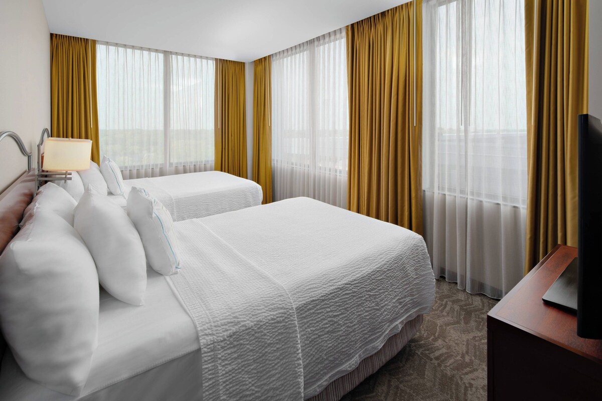 A hotel room with two beds, a TV, and large windows with yellow curtains.
