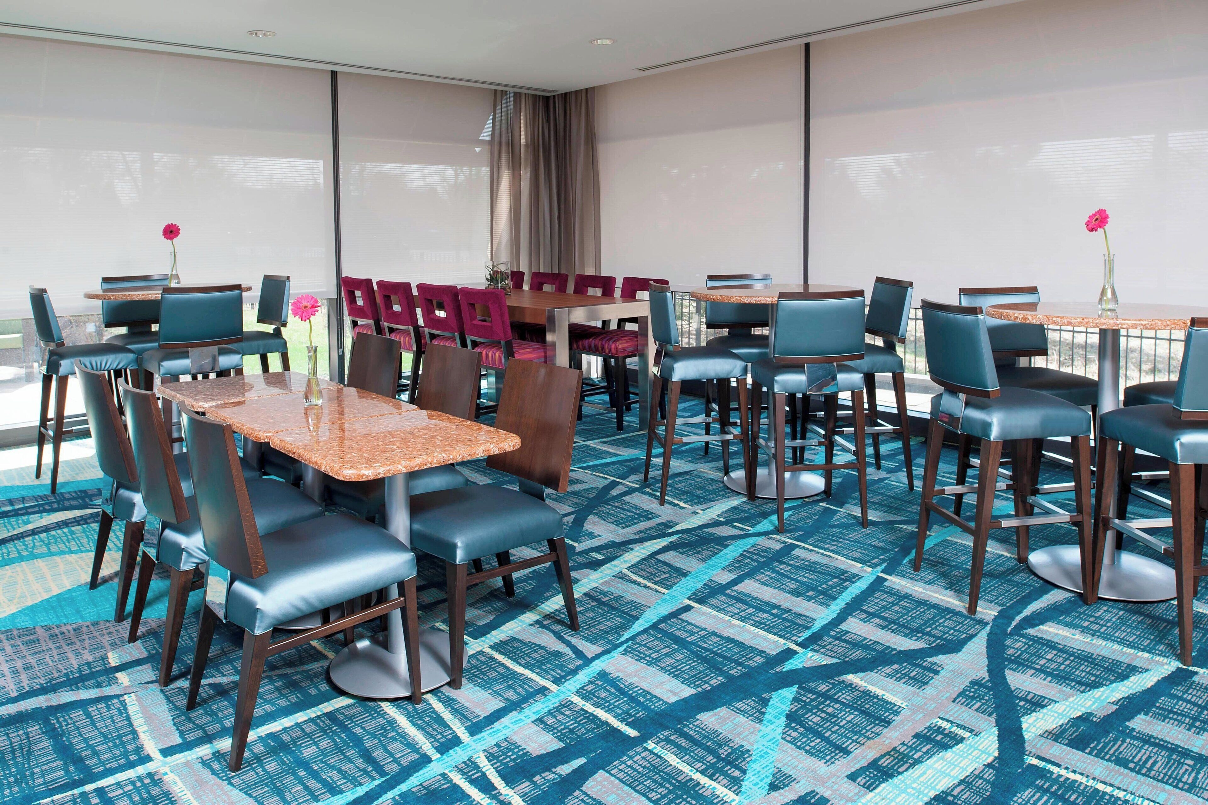 A hotel dining area with multiple tables and chairs, large windows, and a patterned carpet.