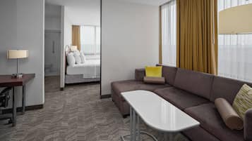 Quarto de hotel com sofá, uma mesinha, cama com cabeceira, luminária e vista para o exterior através de grandes janelas.