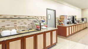 A hotel breakfast area with a long counter, buffet with various food items, a refrigerator, and a coffee machine.